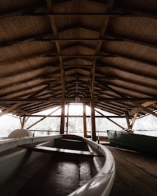 The interior of a wooden boathouse with a high, detailed ceiling and beams made of dark wooden planks. A white rowing boat is positioned inside, with natural light coming through the open sides, revealing snow-covered trees in the background.