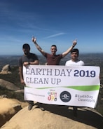 Three individuals are on a rocky hilltop holding a large banner that reads 'Earth Day 2019 Clean Up'. Two of them are raising their arms in a celebratory gesture. The background shows a vast landscape under a clear blue sky.