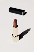 A luxurious lipstick in a sleek gold tube stands upright on a light background. The lipstick color is a rich brown, and a black rectangular case is positioned behind it, casting a soft shadow.
