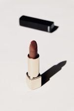 A luxurious lipstick in a sleek gold tube stands upright on a light background. The lipstick color is a rich brown, and a black rectangular case is positioned behind it, casting a soft shadow.