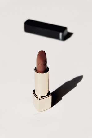 A luxurious lipstick in a sleek gold tube stands upright on a light background. The lipstick color is a rich brown, and a black rectangular case is positioned behind it, casting a soft shadow.