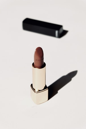 A luxurious lipstick in a sleek gold tube stands upright on a light background. The lipstick color is a rich brown, and a black rectangular case is positioned behind it, casting a soft shadow.