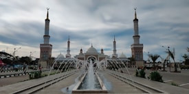A grand mosque with multiple minarets and domes, surrounded by fountains and lush greenery. The pathway leading to the mosque entrance is lined with fountains spraying water into the air, adding a sense of tranquility. The sky is partly cloudy, creating a peaceful and serene atmosphere.
