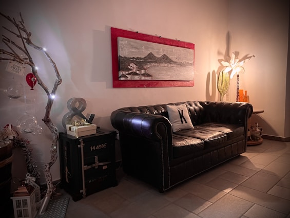 A cozy living room features a dark leather sofa with a white pillow. On the wall above the sofa, there's a framed artwork depicting a landscape with mountains and water. To the left, a decorative branch with lights and hanging ornaments adds a whimsical touch, alongside a metal container with a decorative gift on top. A floor lamp with a white floral shade illuminates the corner, complemented by a tall green vase and orange accent bottles.