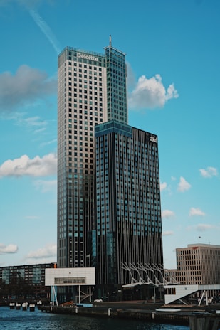 A modern high-rise building with a glass and steel facade, prominently branded with corporate logos near the top. The skyscraper is set against a backdrop of a clear blue sky with scattered clouds. The foreground includes a body of water, part of the urban landscape, and a few smaller buildings.