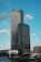 A modern high-rise building with a glass and steel facade, prominently branded with corporate logos near the top. The skyscraper is set against a backdrop of a clear blue sky with scattered clouds. The foreground includes a body of water, part of the urban landscape, and a few smaller buildings.