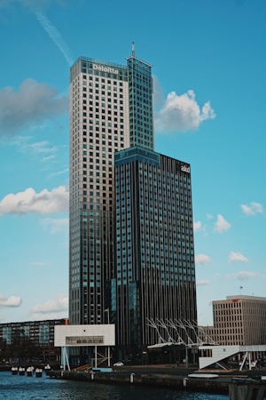 A modern high-rise building with a glass and steel facade, prominently branded with corporate logos near the top. The skyscraper is set against a backdrop of a clear blue sky with scattered clouds. The foreground includes a body of water, part of the urban landscape, and a few smaller buildings.