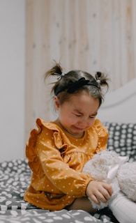 A young child with pigtails is sitting on a polka-dotted bed, wearing an orange polka-dotted dress with ruffled sleeves. The child appears to be upset and is hugging a plush toy tightly.