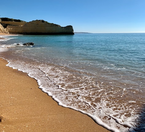 Golden sand beach bordered by gentle waves that wash ashore under a clear blue sky. A rugged cliff extends into the calm, turquoise sea.