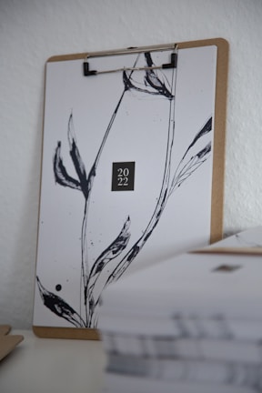 A clipboard is holding a sheet of paper with minimalist black ink design resembling plant stems. The paper prominently displays '2022' in the center. In the foreground, there is a stack of papers that is slightly out of focus.