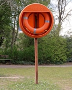 An orange lifebuoy mounted on a wooden post is situated in a grassy area with trees in the background. The word 'LIFEBUOY' is embossed on the outer covering containing the ring.