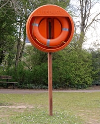An orange lifebuoy mounted on a wooden post is situated in a grassy area with trees in the background. The word 'LIFEBUOY' is embossed on the outer covering containing the ring.