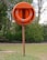 An orange lifebuoy mounted on a wooden post is situated in a grassy area with trees in the background. The word 'LIFEBUOY' is embossed on the outer covering containing the ring.