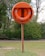 An orange lifebuoy mounted on a wooden post is situated in a grassy area with trees in the background. The word 'LIFEBUOY' is embossed on the outer covering containing the ring.