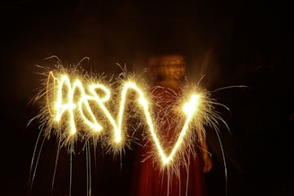 Bright sparklers create streaks of golden light in a dark setting, with a figure partially visible in the background wearing red clothing. The pattern formed by the sparklers resembles a wave or swoosh.