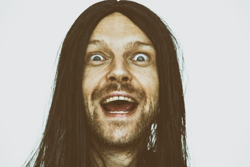 A person with long dark hair appears to be excited or surprised, with wide open eyes and mouth. The facial expression suggests a strong emotion. The background is plain and light-colored.