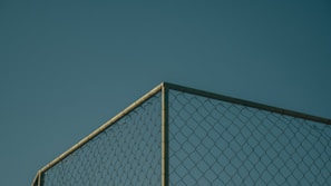A section of a chain-link fence against a clear, deep blue sky. The fence is positioned at a corner, with metal posts visible, casting subtle shadows and highlighting the geometric pattern of the links.