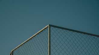 A section of a chain-link fence against a clear, deep blue sky. The fence is positioned at a corner, with metal posts visible, casting subtle shadows and highlighting the geometric pattern of the links.