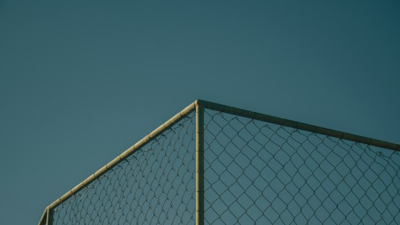 A section of a chain-link fence against a clear, deep blue sky. The fence is positioned at a corner, with metal posts visible, casting subtle shadows and highlighting the geometric pattern of the links.