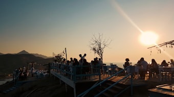 A group of people sitting at tables on an outdoor terrace during sunset. The scene overlooks a mountainous landscape, with clear skies and a prominent sun casting an orange glow. Sparse trees and decorations enhance the setting's tranquil atmosphere.