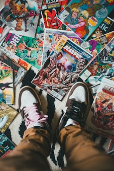 A collection of colorful comic book covers spread across a surface, surrounded by a person's feet wearing mismatched sneakers, one with pink laces and the other with black laces.