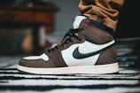 A close-up of a sneaker with a prominent black swoosh logo on the side. The shoe features a combination of dark brown and white leather, with black laces and accents. It is worn with tan pants, and the background includes a blurred piece of furniture and a striped rug.