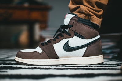 A close-up of a sneaker with a prominent black swoosh logo on the side. The shoe features a combination of dark brown and white leather, with black laces and accents. It is worn with tan pants, and the background includes a blurred piece of furniture and a striped rug.