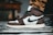 A close-up of a sneaker with a prominent black swoosh logo on the side. The shoe features a combination of dark brown and white leather, with black laces and accents. It is worn with tan pants, and the background includes a blurred piece of furniture and a striped rug.