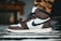 A close-up of a sneaker with a prominent black swoosh logo on the side. The shoe features a combination of dark brown and white leather, with black laces and accents. It is worn with tan pants, and the background includes a blurred piece of furniture and a striped rug.