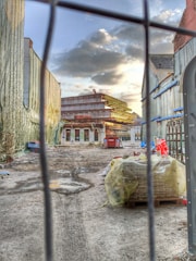 A construction site with a building under construction in the distance, surrounded by scaffolding. The area is littered with construction materials and debris. The foreground shows a dirt ground with puddles, and some materials covered with plastic. The sky above is partly cloudy, with the sun setting, casting an orange glow on the scene.
