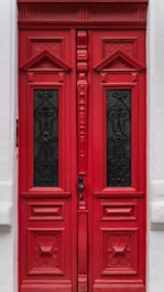 A red wooden double door with ornate, carved designs and two rectangular black panels featuring intricate wrought iron patterns. The door is set within a white painted wall, with small decorative elements framing it.