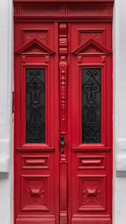 A red wooden double door with ornate, carved designs and two rectangular black panels featuring intricate wrought iron patterns. The door is set within a white painted wall, with small decorative elements framing it.