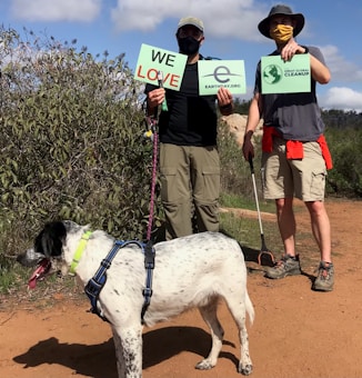 Two individuals are outdoors, holding signs related to Earth Day and environmental cleanup efforts. They are accompanied by a large dog on a leash. The scene is set on a dirt path surrounded by greenery, and both individuals are wearing hats and face masks, suggesting sunny weather and safety precautions.