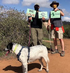 Two individuals are outdoors, holding signs related to Earth Day and environmental cleanup efforts. They are accompanied by a large dog on a leash. The scene is set on a dirt path surrounded by greenery, and both individuals are wearing hats and face masks, suggesting sunny weather and safety precautions.