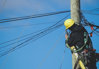 man in black jacket and yellow hard hat climbing on brown wooden post during daytime