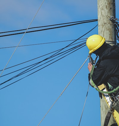 man in black jacket and yellow hard hat climbing on brown wooden post during daytime