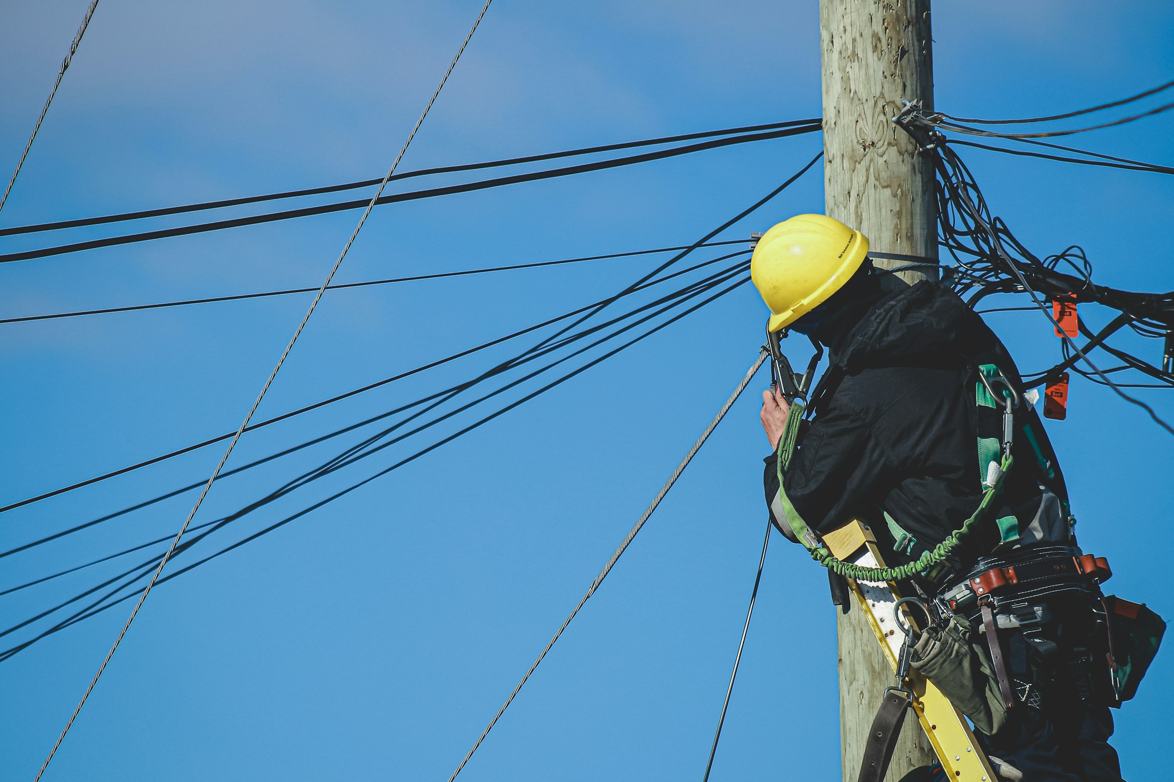 A lineworker in a yellow hard hat and climbing harness works at the top of a wooden distribution pole, surrounded by overhead conductors against a clear blue sky.