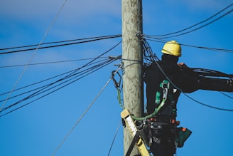 Worker wearing Turtel Armor harness and helmet while climbing a telecommunications tower against a clear blue sky.