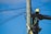 A worker wearing a hard hat and safety harness is climbing a wooden utility pole, surrounded by numerous cables against a clear blue sky.
