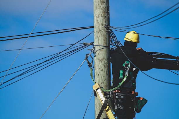 Worker wearing Turtel Armor harness and helmet while climbing a telecommunications tower against a clear blue sky.