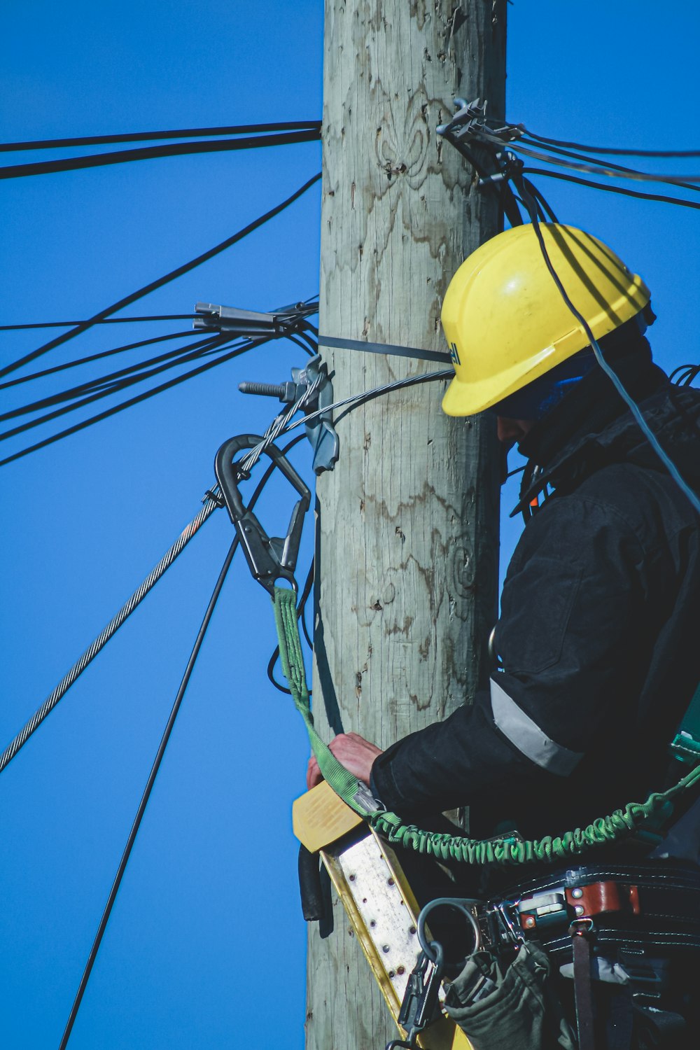 man in black jacket and yellow hard hat climbing on brown wooden post during daytime