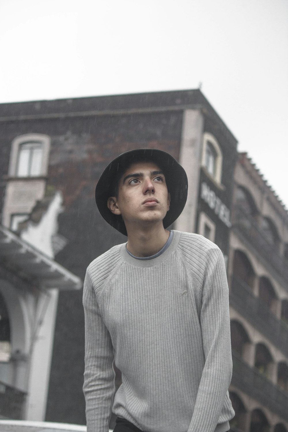 man in gray crew neck sweater wearing black hat standing near brown concrete building during daytime