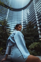 A person wearing a light blue outfit, seated with a modern architectural backdrop. The tall, curved building features numerous windowed floors and is surrounded by greenery, creating a futuristic and urban atmosphere.