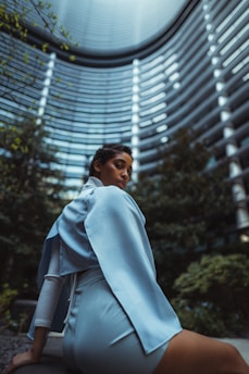 A person wearing a light blue outfit, seated with a modern architectural backdrop. The tall, curved building features numerous windowed floors and is surrounded by greenery, creating a futuristic and urban atmosphere.