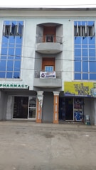 A three-story building featuring large blue reflective windows and a central balcony. The ground floor has two storefronts: a pharmacy and another shop with a yellow sign that has text and graphics. The exterior walls are painted a grayish color, and two columns support the balcony. The street in front appears empty.