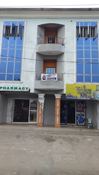 A three-story building featuring large blue reflective windows and a central balcony. The ground floor has two storefronts: a pharmacy and another shop with a yellow sign that has text and graphics. The exterior walls are painted a grayish color, and two columns support the balcony. The street in front appears empty.