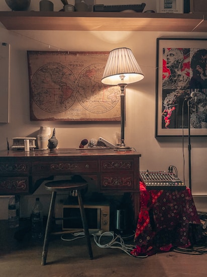 A cozy, dimly-lit room showcasing an ornate wooden desk with intricate patterns. On the desk are decorative items, including a small skull sculpture and a lamp casting a warm glow. Behind the desk, a vintage world map hangs next to a modern framed artwork depicting faces with bold red accents. A mixer and other electronic equipment are on another surface, partially covered with a red-patterned cloth. A stool is tucked under the desk, and various cords are visible around the floor.