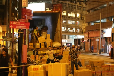 Several people are unloading boxes from a truck onto the street in an urban environment. The scene appears to be bustling with activity, as there are numerous packages and several individuals working together. The buildings in the background suggest a commercial area, and the lighting indicates that it might be evening.