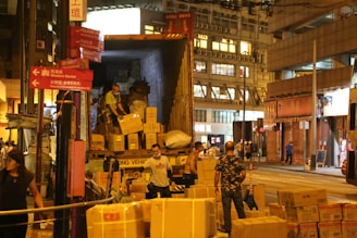 Several people are unloading boxes from a truck onto the street in an urban environment. The scene appears to be bustling with activity, as there are numerous packages and several individuals working together. The buildings in the background suggest a commercial area, and the lighting indicates that it might be evening.