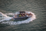 A motorboat speeds through the water, creating wake patterns behind it. Several people are onboard, some sitting and others standing, likely enjoying a leisure activity. The boat has a sleek design and appears to be modern.